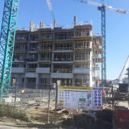 Wielopiętrowy budynek w trakcie budowy, widoczne szalunki stropowe i zbrojenia, na pierwszym planie tablica informacyjna 'Strefa Bezpieczeństwa - Plac Budowy', w tle żurawie budowlane i pompa...