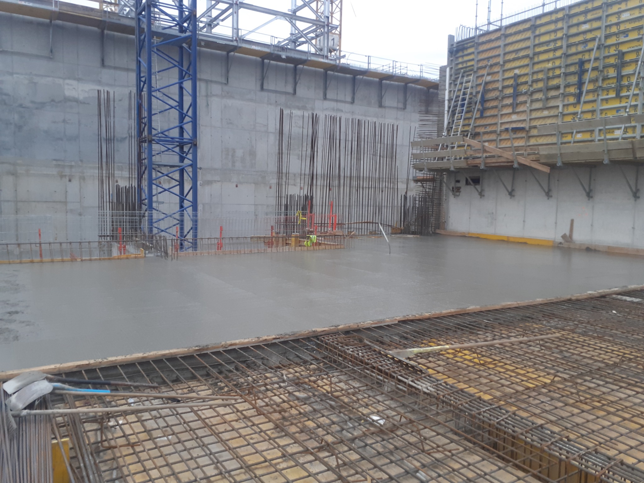 Świeżo wylana, gładka wylewka betonowa na budowie, widoczna siatka zbrojeniowa w pierwszym planie i pionowe pręty zbrojeniowe wystające z betonu, żółte szalunki na ścianach w tle i niebieski żuraw...