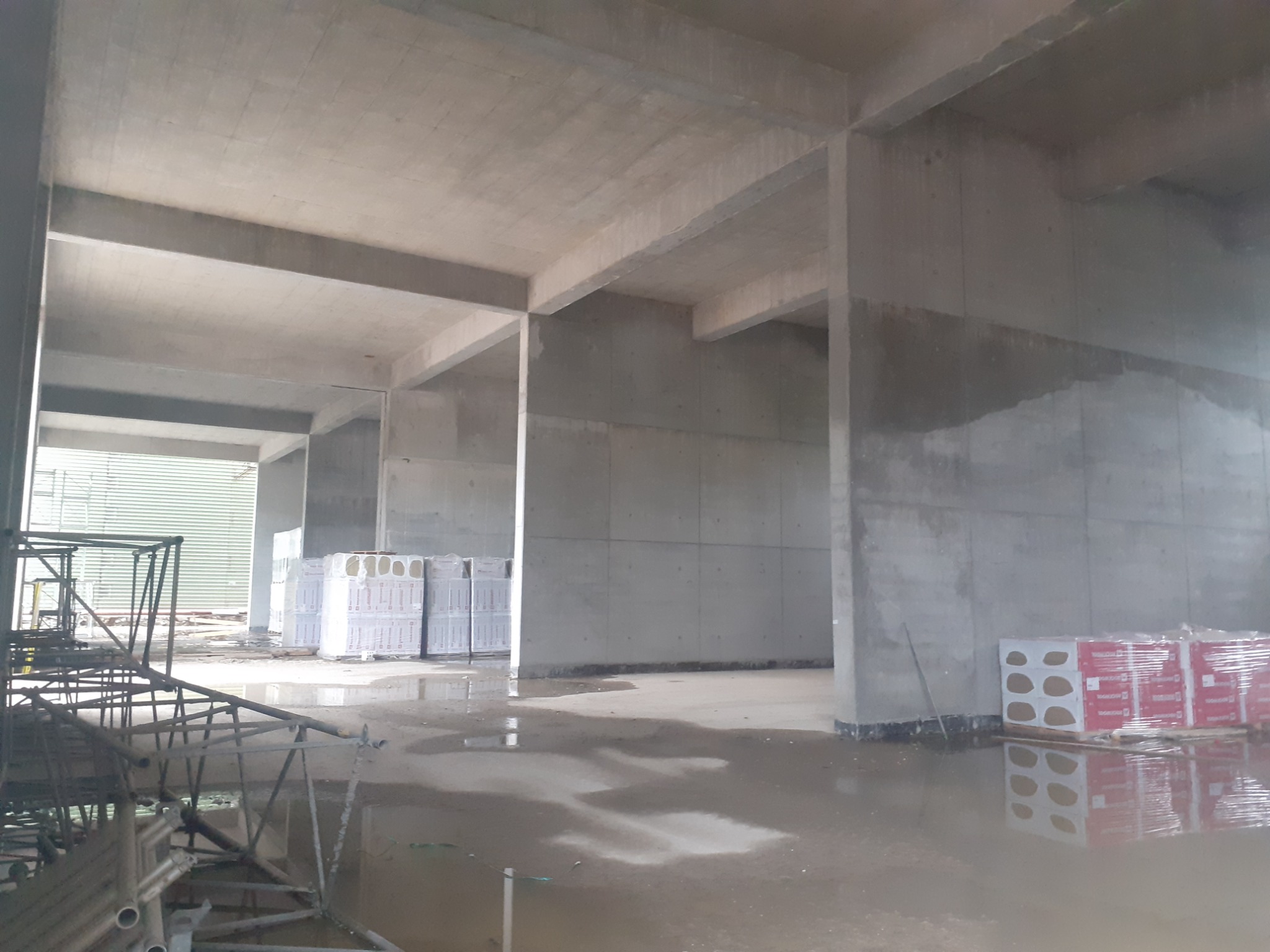 Wnętrze surowego budynku w trakcie budowy z widocznymi betonowymi filarami, stropem, mokrą posadzką, rusztowaniami i paletami materiałów budowlanych w folii.