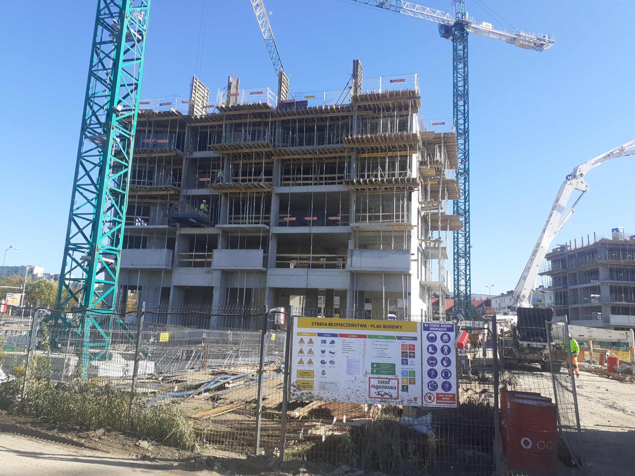 Wielopiętrowy budynek w trakcie budowy, widoczne szalunki stropowe i zbrojenia, na pierwszym planie tablica informacyjna 'Strefa Bezpieczeństwa - Plac Budowy', w tle żurawie budowlane i pompa...