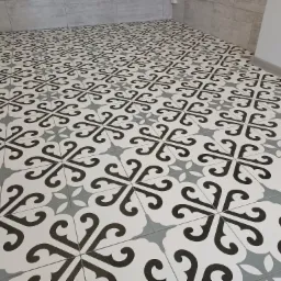 Wzorzysta podłoga z płytek ceramicznych w odcieniach bieli, szarości i czerni, ułożona w regularny deseń, widoczna przy ścianie wykończonej szarymi płytami.