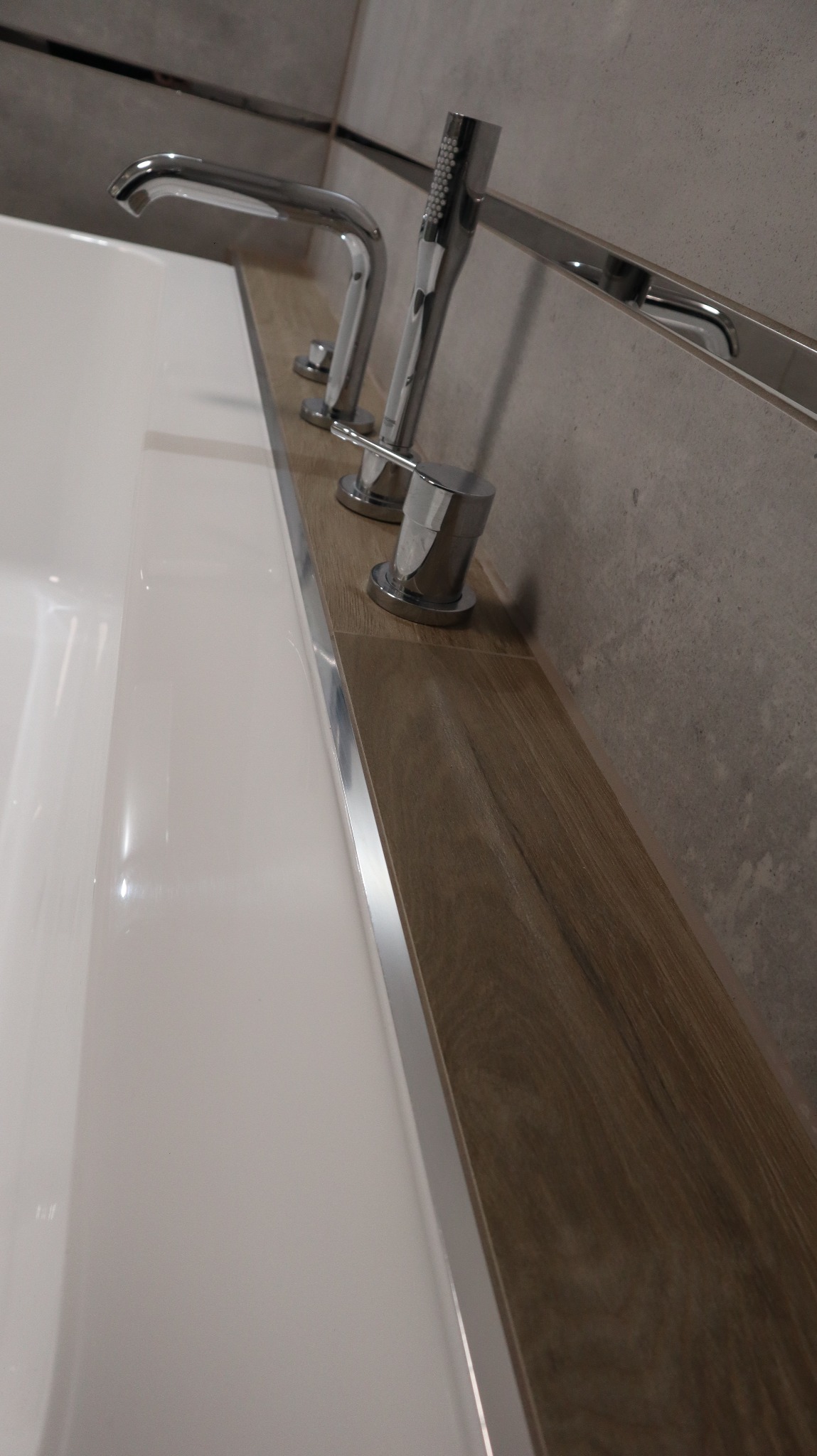 Bateria wannowa z chromowanymi elementami, zamontowana na drewnopodobnej półce przy wannie, z szarą płytką w tle.