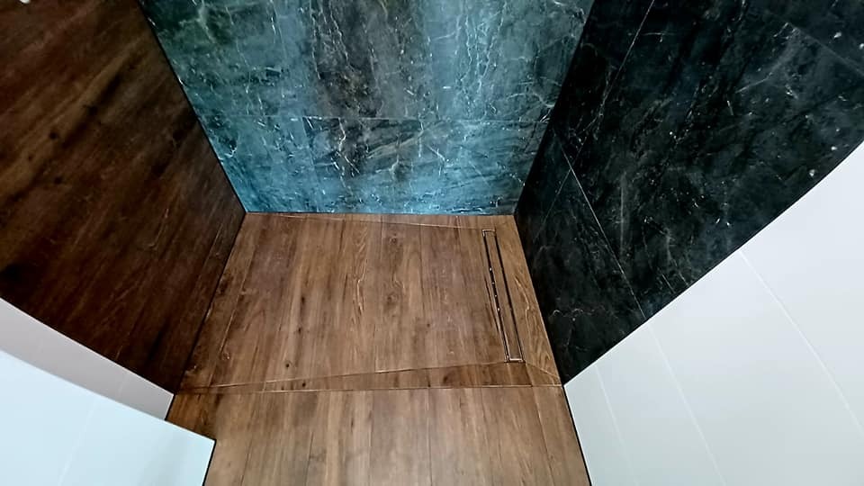 Nowoczesna kabina prysznicowa walk-in z odpływem liniowym, wykończona płytkami imitującymi drewno na podłodze i marmuropodobnymi płytkami w odcieniach szarości na ścianach.