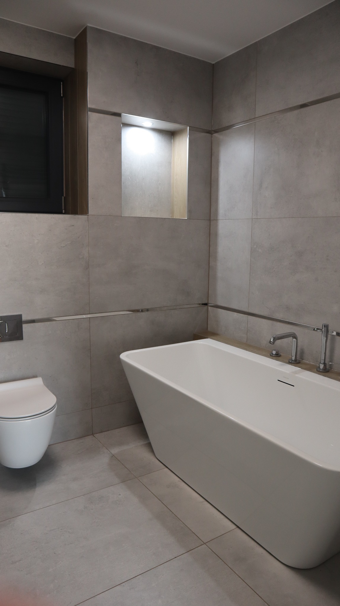 Nowoczesna łazienka z szarymi, wielkoformatowymi płytkami, białą wanną wolnostojącą i podświetlaną wnęką w ścianie.