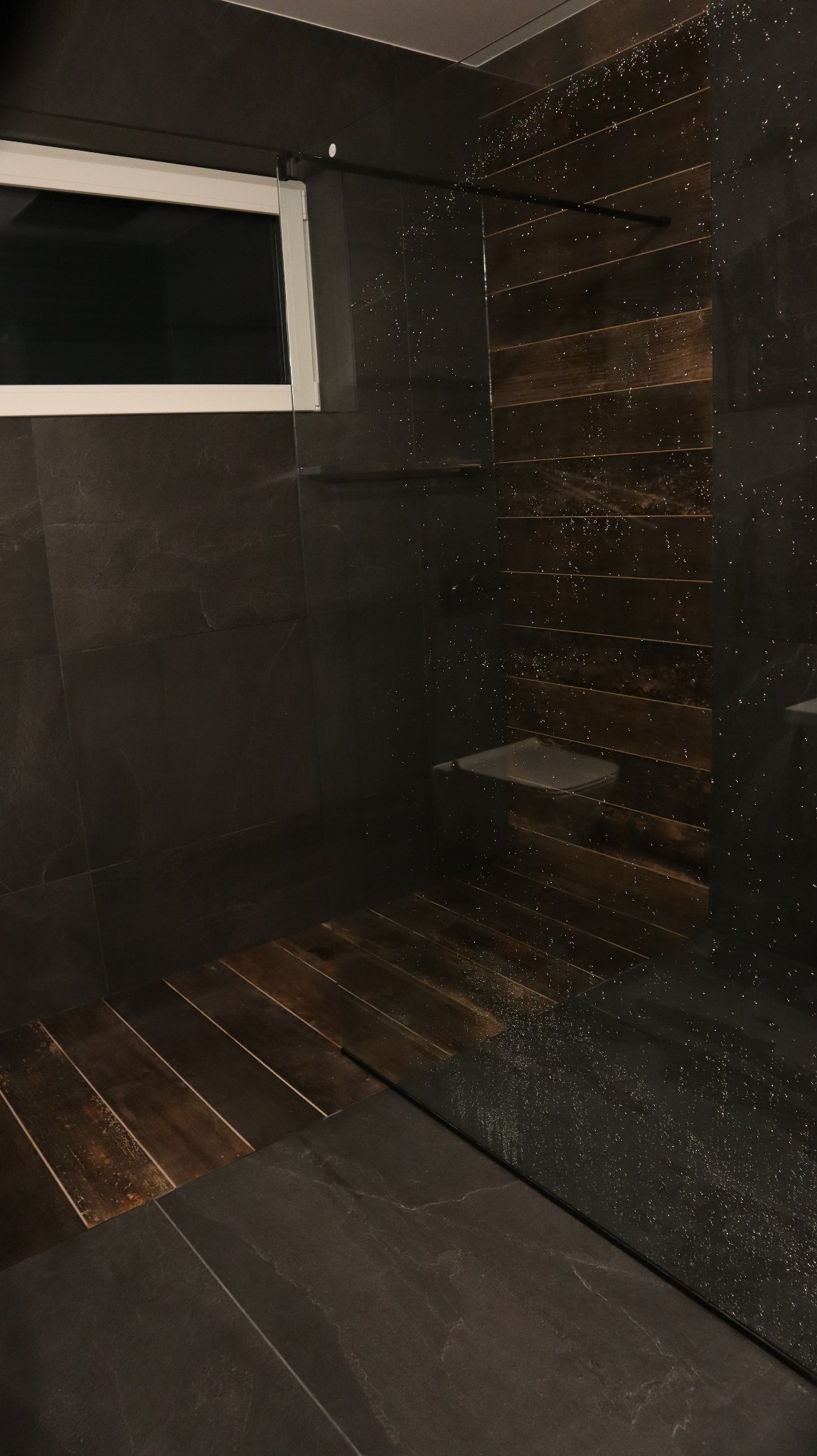 Nowoczesna kabina prysznicowa z ciemnymi płytkami imitującymi kamień i drewnianą podłogą, widoczne krople wody na szklanej ściance.