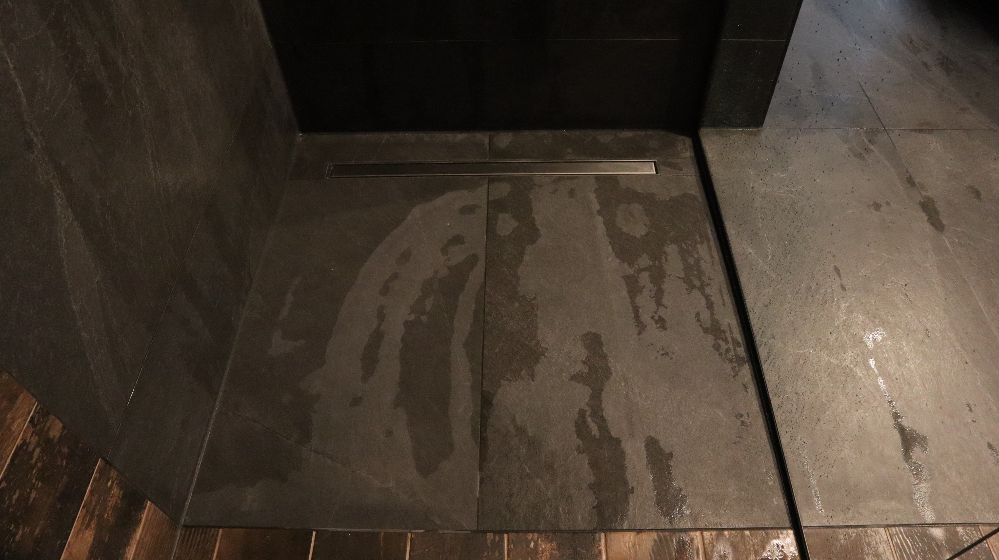 Ciemne płytki podłogowe i ścienne w łazience, z odpływem liniowym. Widoczne ślady wody na powierzchni płytek. Dolna część ściany wykończona drewnopodobnymi płytkami.