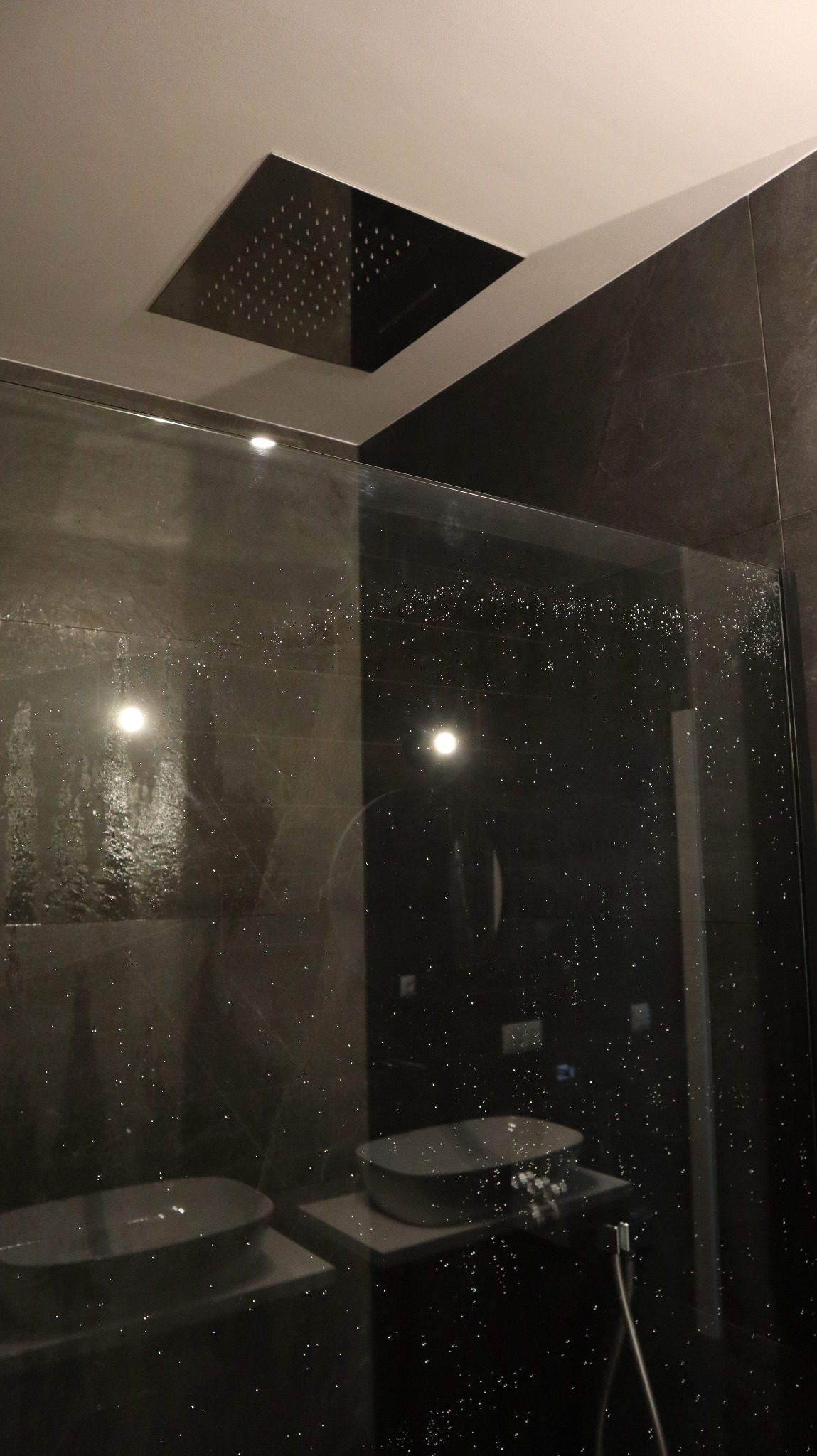 Nowoczesna łazienka z deszczownicą w suficie, szklaną kabiną prysznicową i umywalkami na blacie, wykończona ciemnymi płytkami imitującymi kamień.