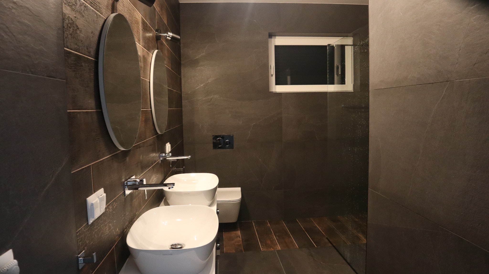 Nowoczesna łazienka z ciemnymi, marmurkowymi płytkami, dwiema owalnymi umywalkami nablatowymi, okrągłymi lustrami, drewnianą podłogą i oknem z białymi ramami.