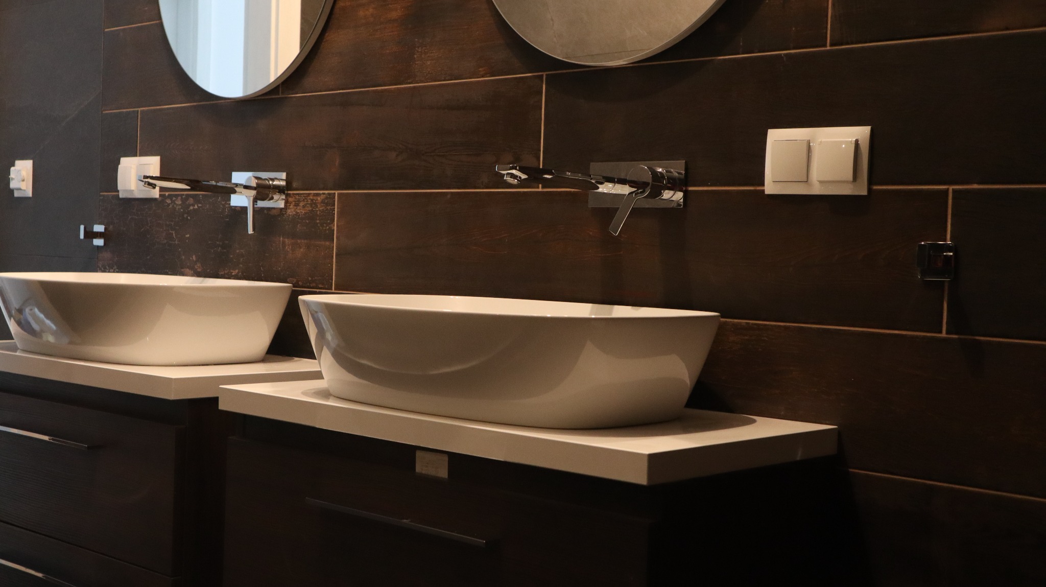 Nowoczesna łazienka z dwoma białymi umywalkami nablatowymi o owalnym kształcie, zamontowanymi na ciemnych szafkach z szufladami. Ściany wyłożone ciemnobrązowymi, poziomymi płytkami imitującymi...