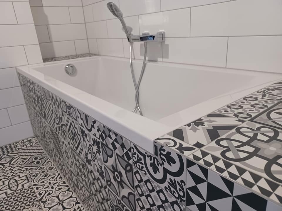 Biała wanna obudowana wzorzystymi, czarno-białymi płytkami ceramicznymi, dopasowanymi do płytek podłogowych w łazience w Lublińcu. Widoczne białe płytki na ścianie i chromowana bateria.
