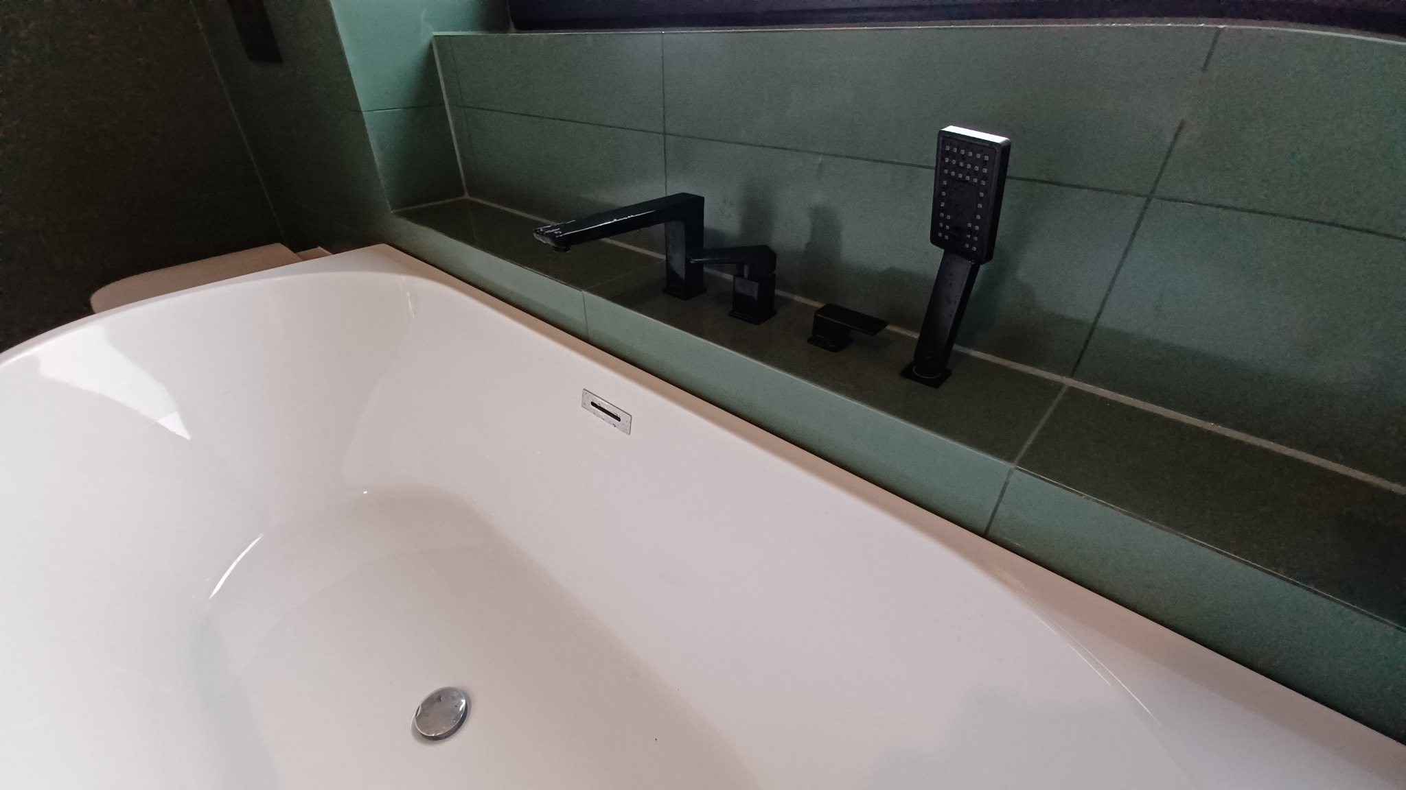 Nowoczesna, biała wanna z czarną armaturą i deszczownicą na tle zielonych płytek ceramicznych.