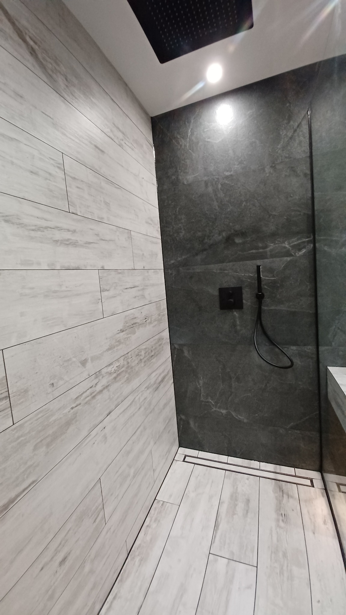 Nowoczesna kabina prysznicowa z szarymi płytkami imitującymi marmur na ścianach i podłodze, wyposażona w deszczownicę i czarną armaturę podtynkową.