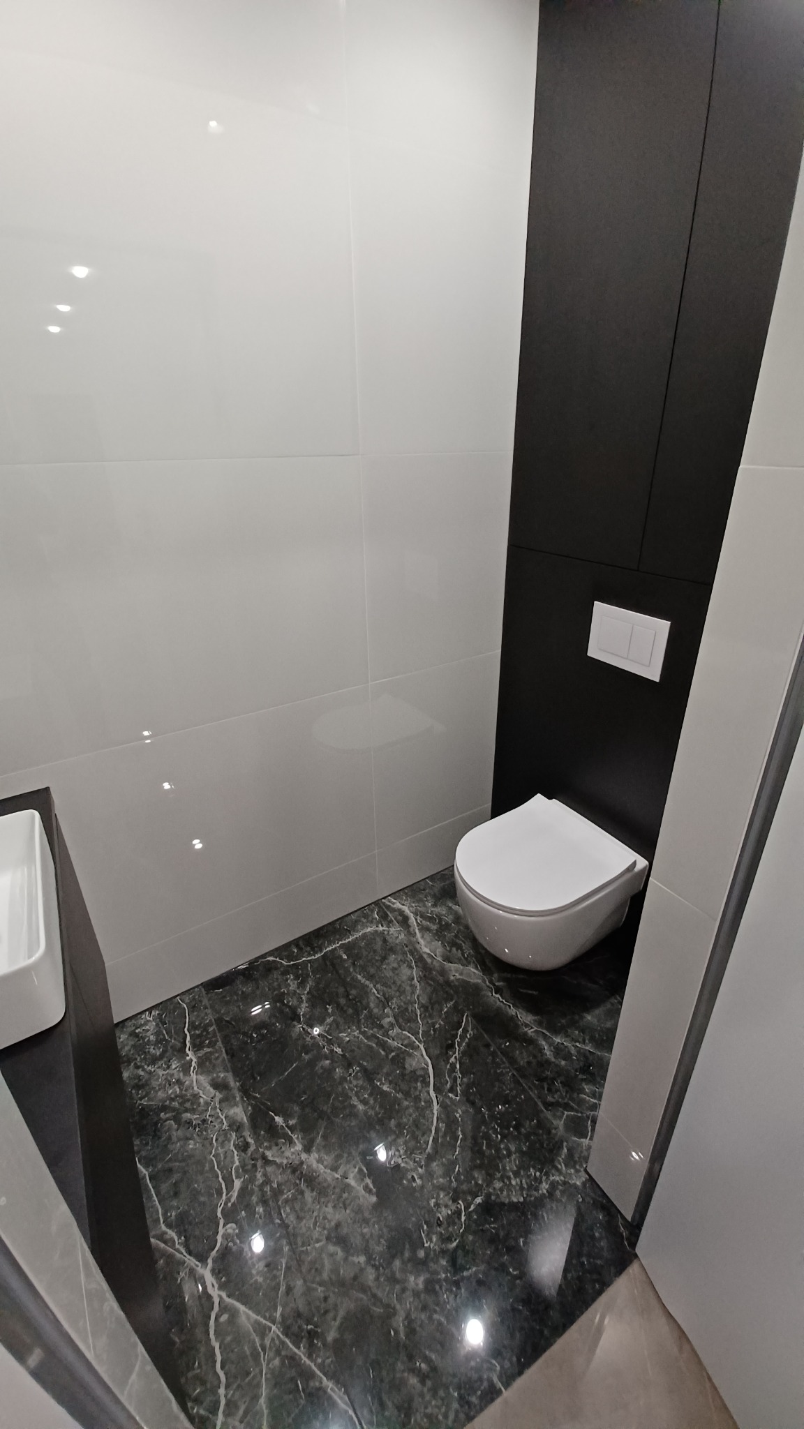 Nowoczesna łazienka z białymi płytkami ściennymi, czarną zabudową geberit i podłogą z płytek imitujących czarny marmur z białymi żyłami, widok z góry.