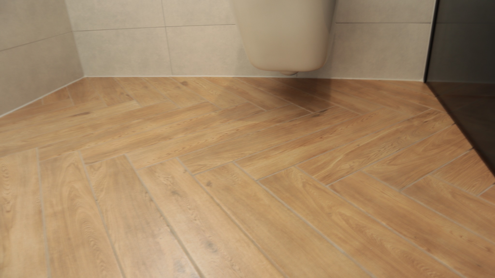 Ułożona w jodełkę podłoga z płytek imitujących drewno w łazience z podwieszaną toaletą i jasnymi płytkami ściennymi.