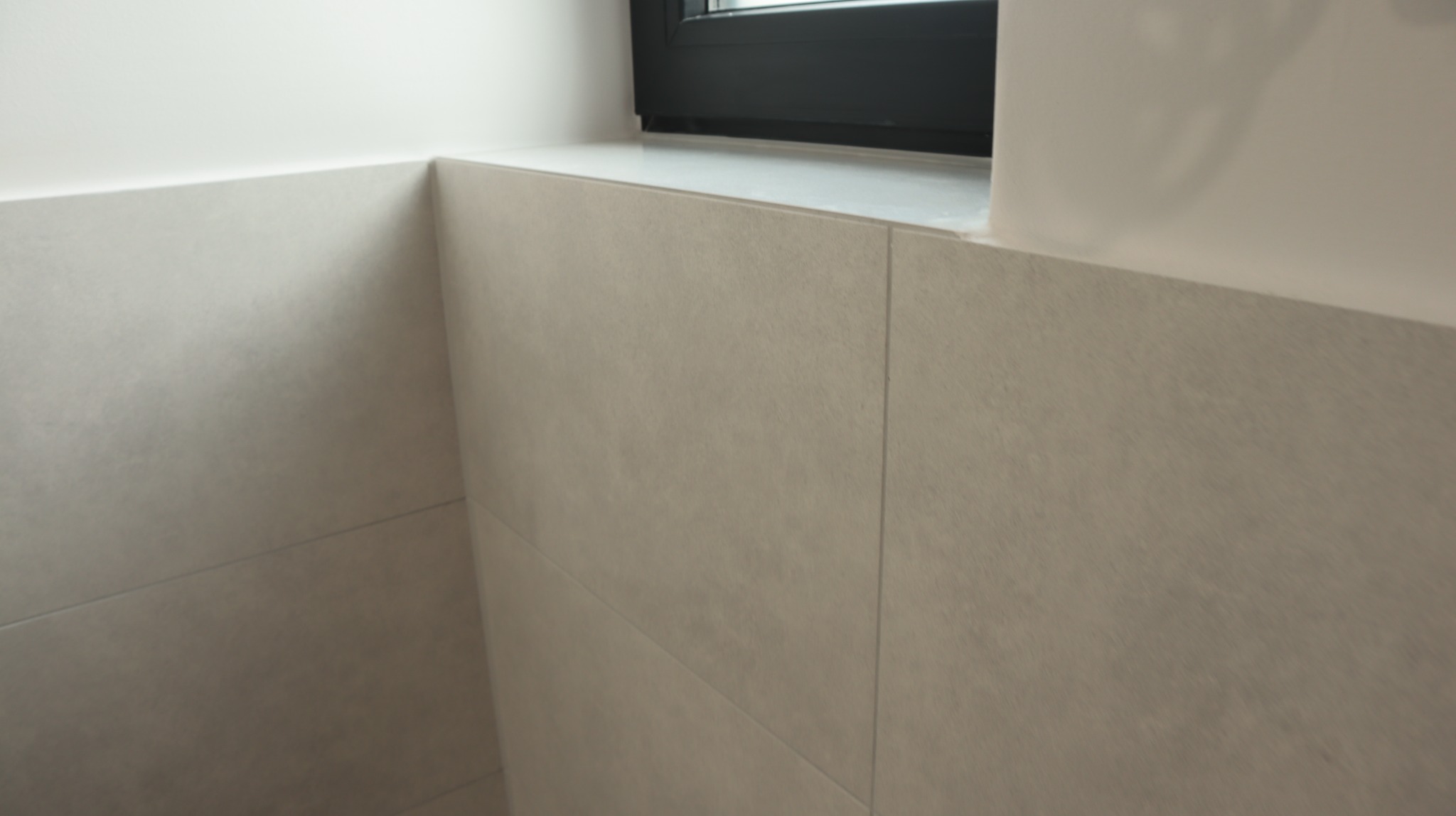 Jasne, matowe płytki ceramiczne ułożone na ścianie w narożniku przy oknie, widoczne fugi i delikatna tekstura płytek.