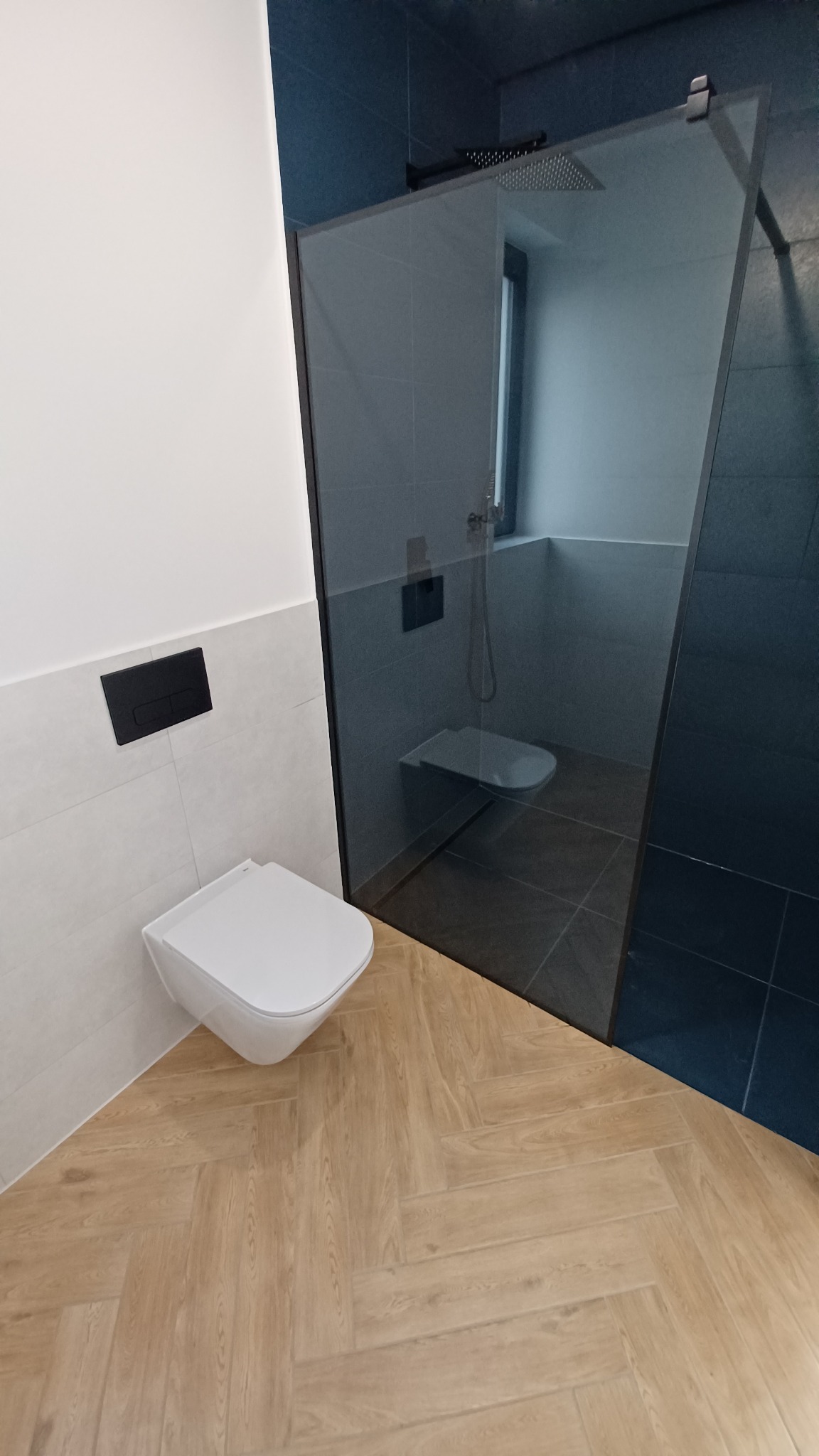 Nowoczesna łazienka z kabiną typu walk-in, wiszącą toaletą z czarnym przyciskiem spłukującym i podłogą z paneli ułożonych w jodełkę.