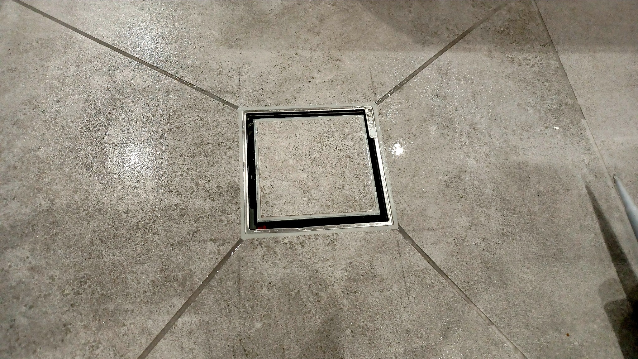 Szara, matowa płytka podłogowa z kwadratowym odpływem liniowym z czarną ramką, ułożona zbieżnie do środka odpływu, widoczne fugi krzyżujące się w narożnikach odpływu.