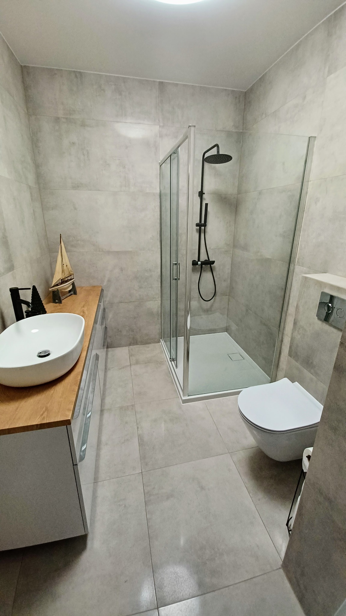 Nowoczesna łazienka z szarymi płytkami, kabiną prysznicową typu walk-in z czarną armaturą, umywalką nablatową na drewnianym blacie i dekoracyjnym modelem żaglówki.