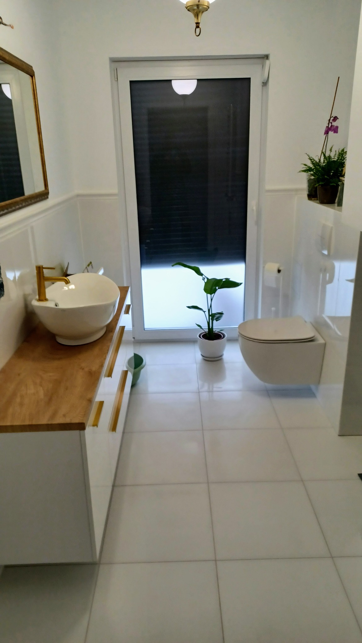 Nowoczesna łazienka w jasnych barwach z drewnianym blatem pod umywalką, złotą armaturą, toaletą podwieszaną i roślinami doniczkowymi.