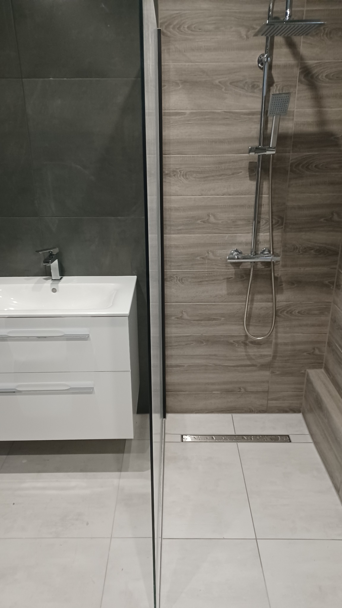 Wykończona łazienka z kabiną prysznicową typu walk-in, drewnopodobnymi płytkami na ścianach, białą szafką z umywalką i szarą ścianą z płytek.
