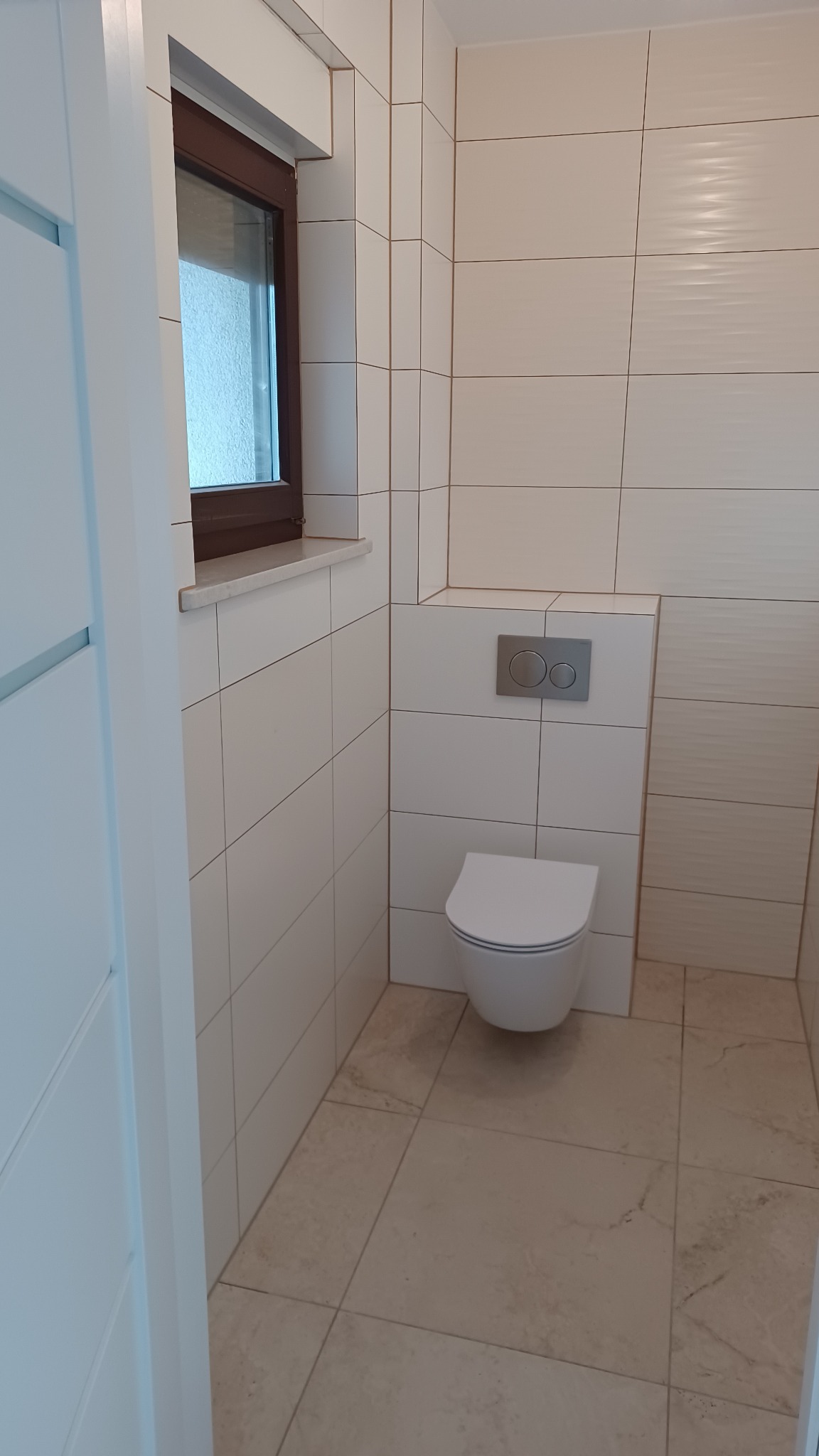 Nowoczesna łazienka z podwieszanym WC, beżowymi płytkami na podłodze i ścianach, oraz oknem z brązową ramą