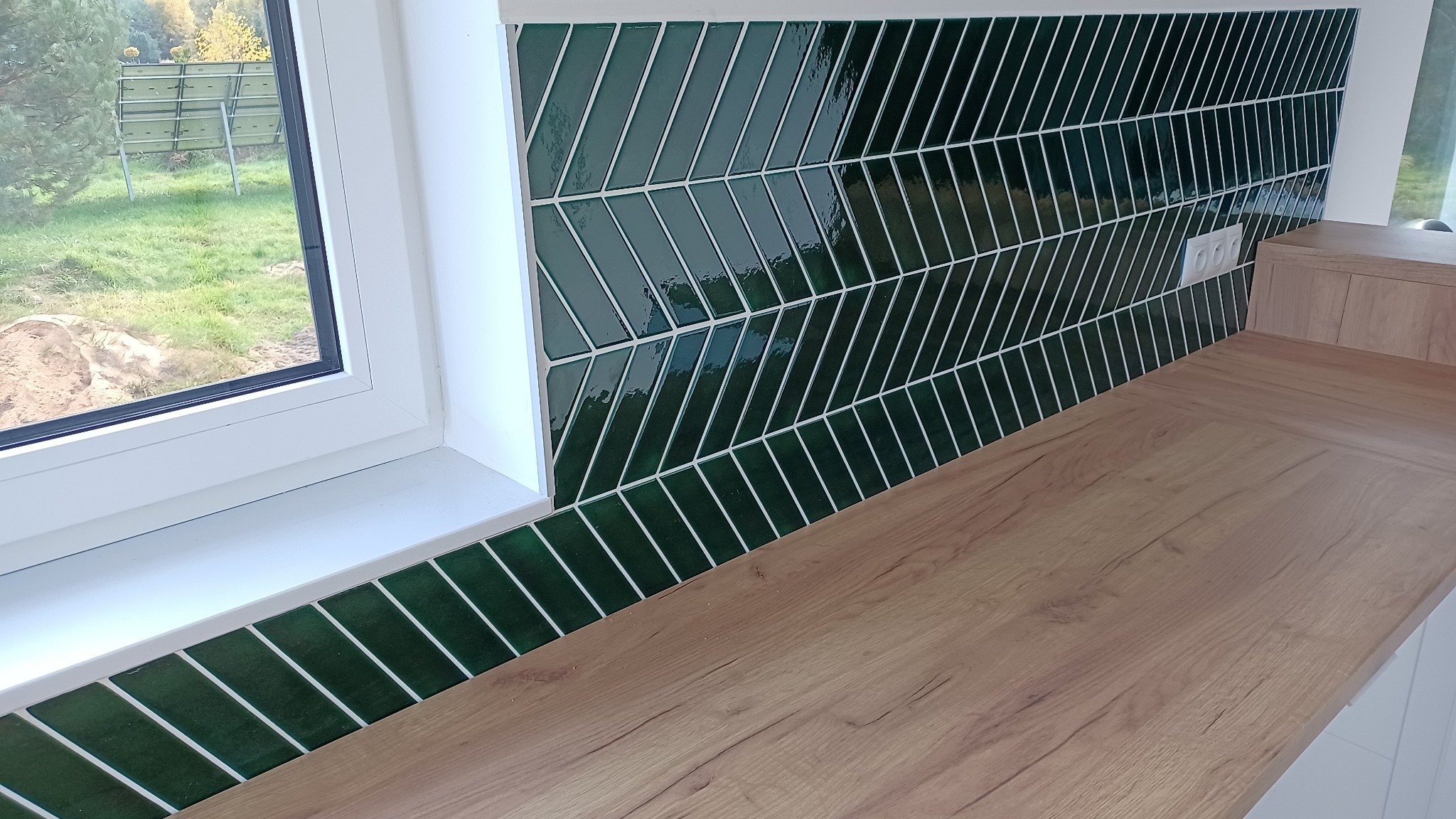 Fragment kuchni z blatem imitującym drewno i ścianą wyłożoną ciemnozielonymi płytkami ułożonymi we wzór jodełki, widoczne gniazdko elektryczne i okno z widokiem na zewnątrz.