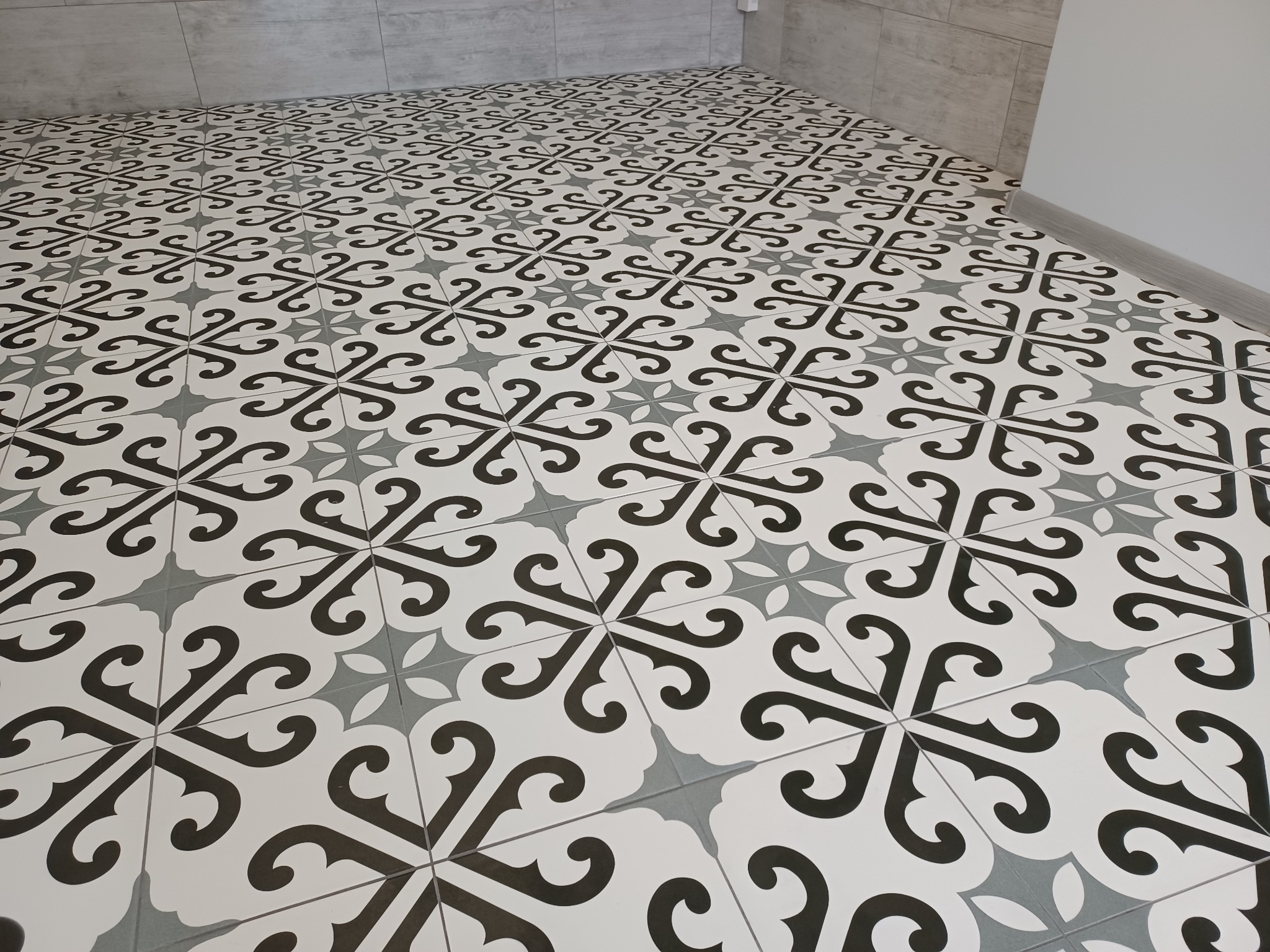 Wzorzysta podłoga z płytek ceramicznych w odcieniach bieli, szarości i czerni, ułożona w regularny deseń, widoczna przy ścianie wykończonej szarymi płytami.