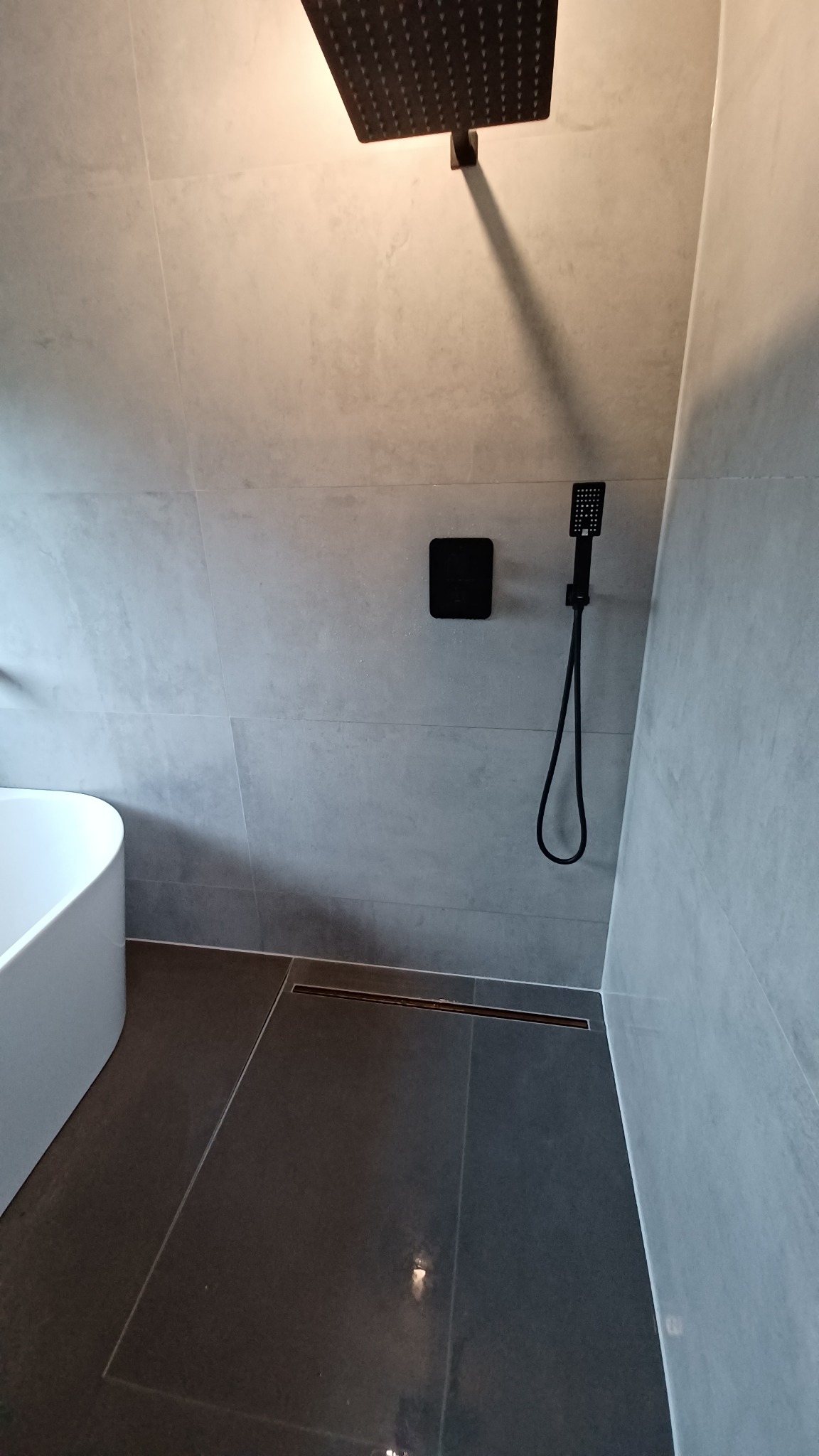 Nowoczesna łazienka z szarymi płytkami, czarną armaturą i odpływem liniowym, widoczny fragment białej wanny.