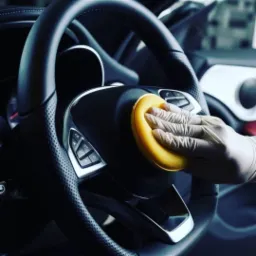 Czyszczenie skórzanej kierownicy samochodu żółtą gąbką przez osobę w rękawiczkach ochronnych.