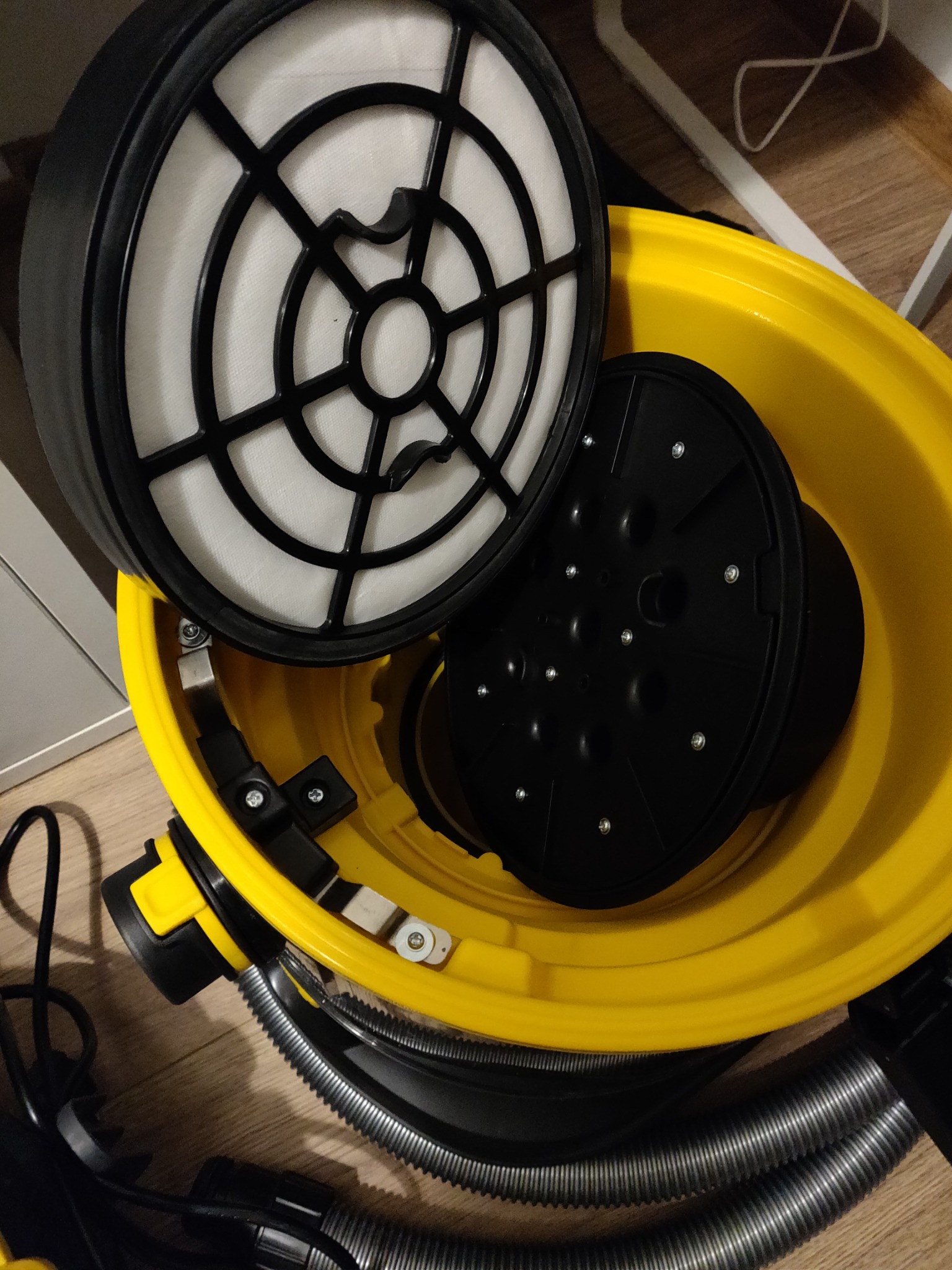 Wnętrze żółtego odkurzacza przemysłowego z widocznym filtrem HEPA i elementami mocującymi, czarny wąż karbowany na podłodze.