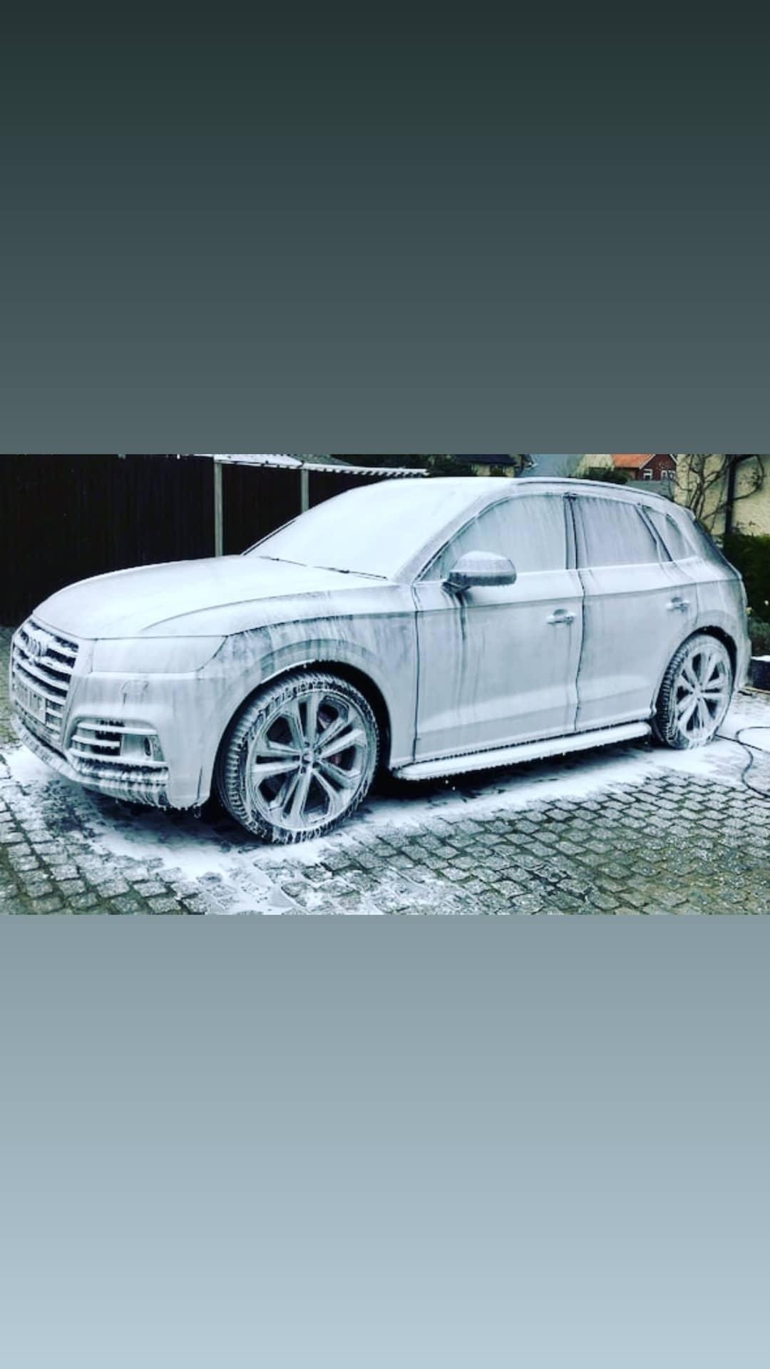 Szary SUV marki Audi pokryty grubą warstwą piany aktywnej podczas mycia na zewnątrz, z widoczną kostką brukową i fragmentem ogrodzenia w tle.