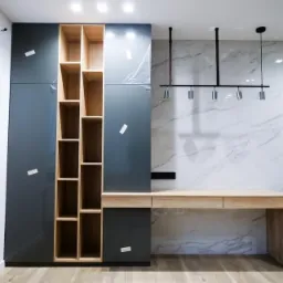 Szare szafy z drewnianymi półkami i biurko z jasnego drewna na tle marmurowej ściany, widoczne folie ochronne na frontach szafek, nowoczesne oświetlenie sufitowe.