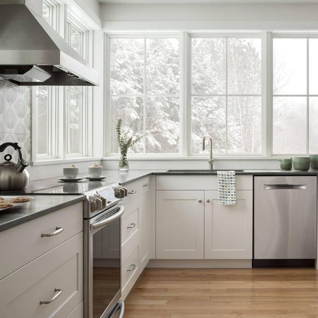 Jasna kuchnia z białymi szafkami i czarnym blatem, widok na zaśnieżony las przez duże okno, drewniana podłoga, srebrny czajnik na płycie grzewczej.