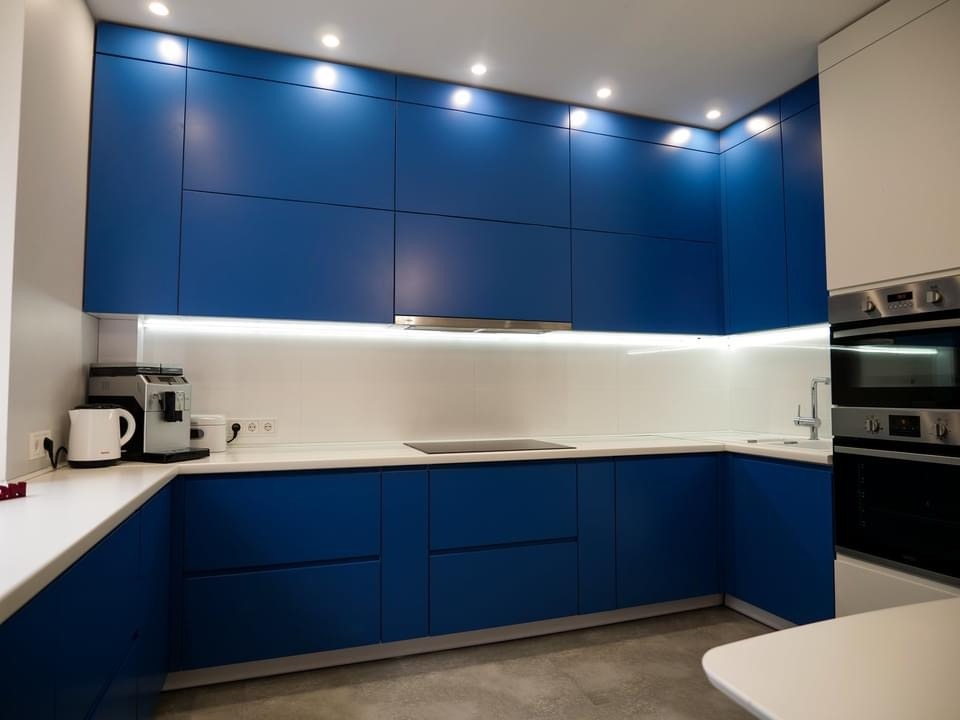 Nowoczesna kuchnia na wymiar z niebieskimi, gładkimi frontami szafek, białym blatem i oświetleniem LED podszafkowym, widoczny ekspres do kawy i piekarnik.