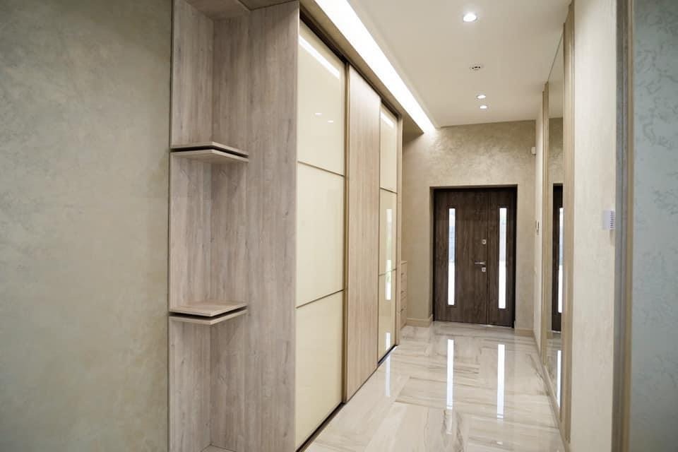 Wnętrze przedpokoju z zabudową szafy przesuwnej z półkami, wykończonej w odcieniach beżu i drewna, z widoczną teksturą ścian i marmurową posadzką.