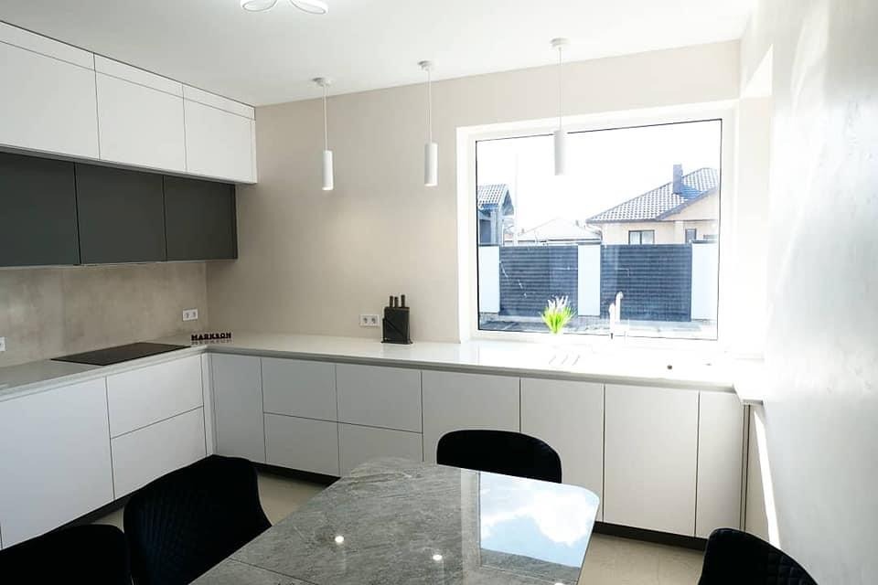 Nowoczesna kuchnia z białymi szafkami dolnymi i szarymi górnymi, minimalistyczne lampy wiszące, blat z czarnego kamienia i stół z szarym blatem.