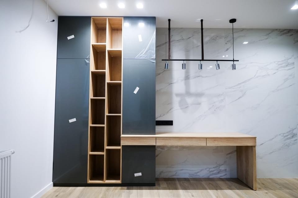 Szare szafy z drewnianymi półkami i biurko z jasnego drewna na tle marmurowej ściany, widoczne folie ochronne na frontach szafek, nowoczesne oświetlenie sufitowe.