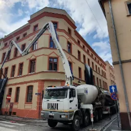 Betonomieszarka z wysięgnikiem podająca beton na elewację kamienicy, zabezpieczoną czarną folią.
