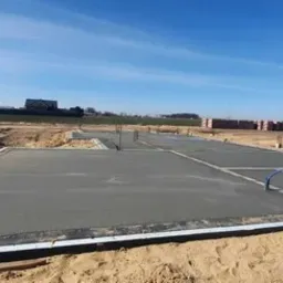 Wylany fundament betonowy na placu budowy, widoczne wystające pręty zbrojeniowe, w tle cegły, samochód i zabudowa jednorodzinna.