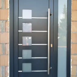 Nowoczesne, grafitowe drzwi wejściowe z czterema kwadratowymi przeszkleniami i długim, pionowym uchwytem, zamontowane w surowym murze z widoczną pianką montażową.