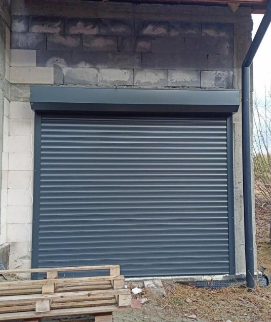 Zamknięta, szara roleta zewnętrzna na surowej ścianie budynku z bloków, z drewnianą paletą u dołu.