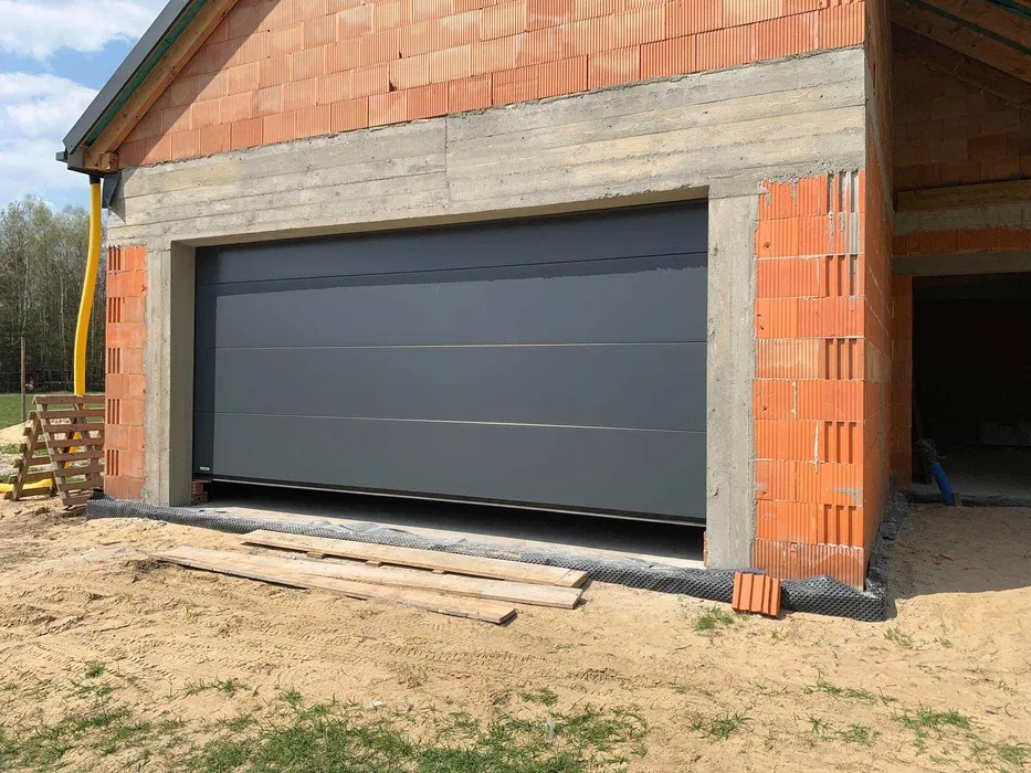 Szara segmentowa konstrukcja w trakcie montażu, widoczne cegły i betonowe obramowanie w surowym stanie, teren wokół wyrównany piaskiem.