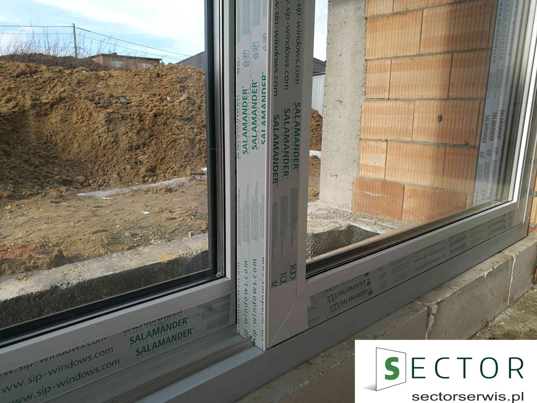 Nowo zamontowane okno aluminiowe marki Salamander w surowym stanie, widok z zewnątrz na plac budowy z ziemią i ceglanym murem.
