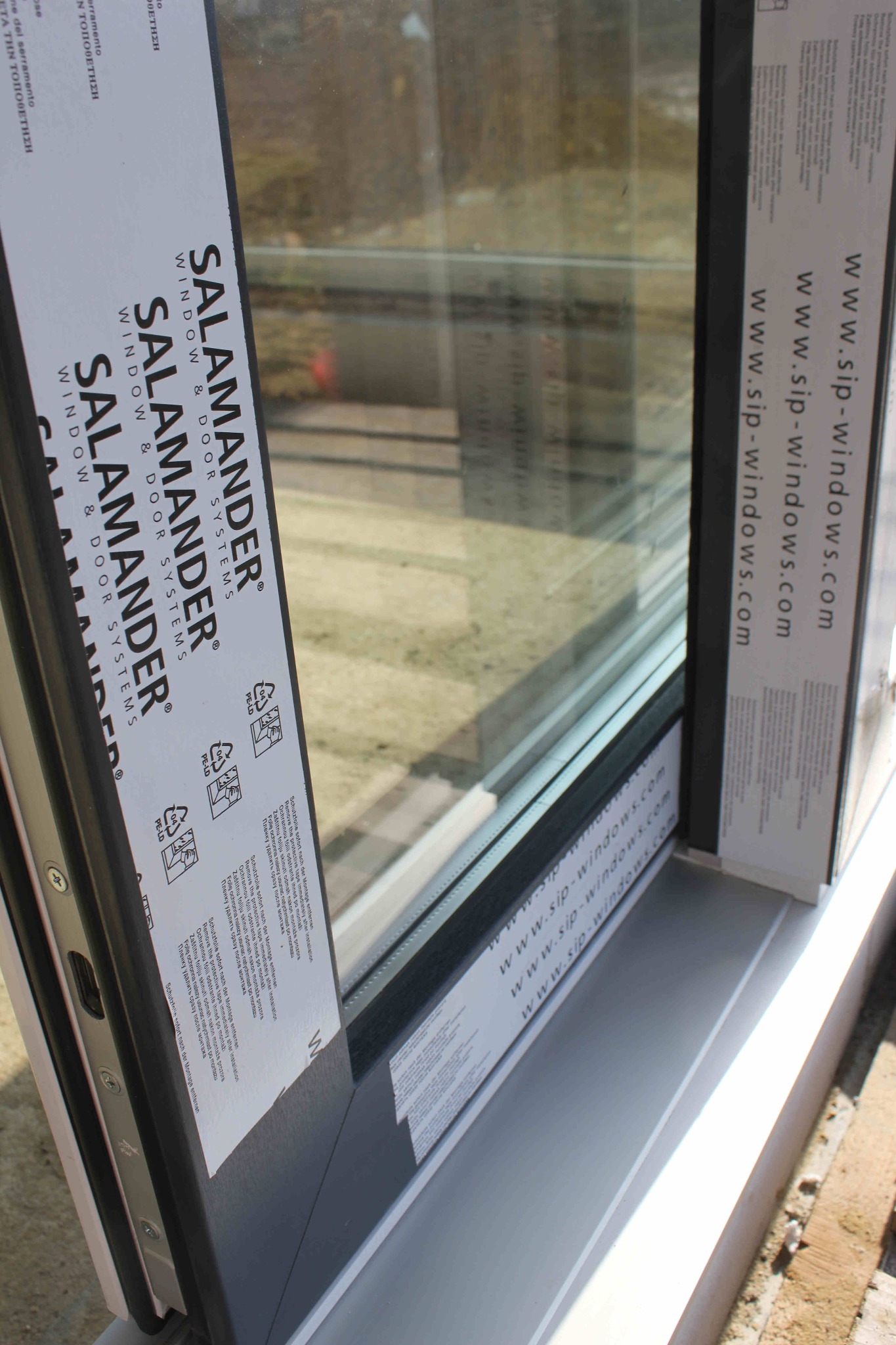 Fragment nowo zamontowanego okna aluminiowego marki Salamander z naklejonymi foliami ochronnymi i informacjami o producencie, widoczne śruby montażowe.