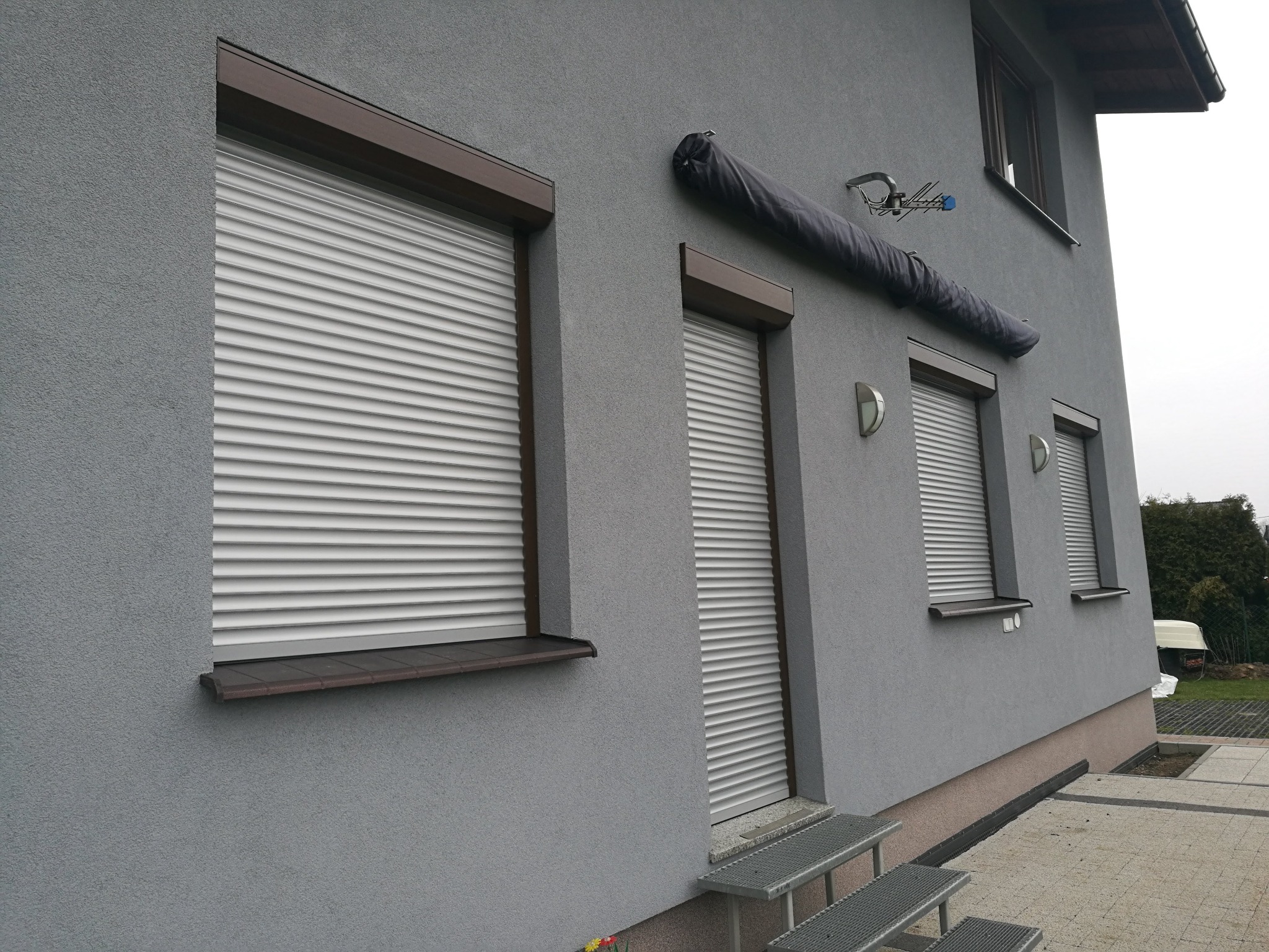 Szara elewacja domu z trzema oknami zabezpieczonymi roletami zewnętrznymi, zadaszenie nad oknem na pierwszym piętrze, schody wejściowe z metalowej kraty.