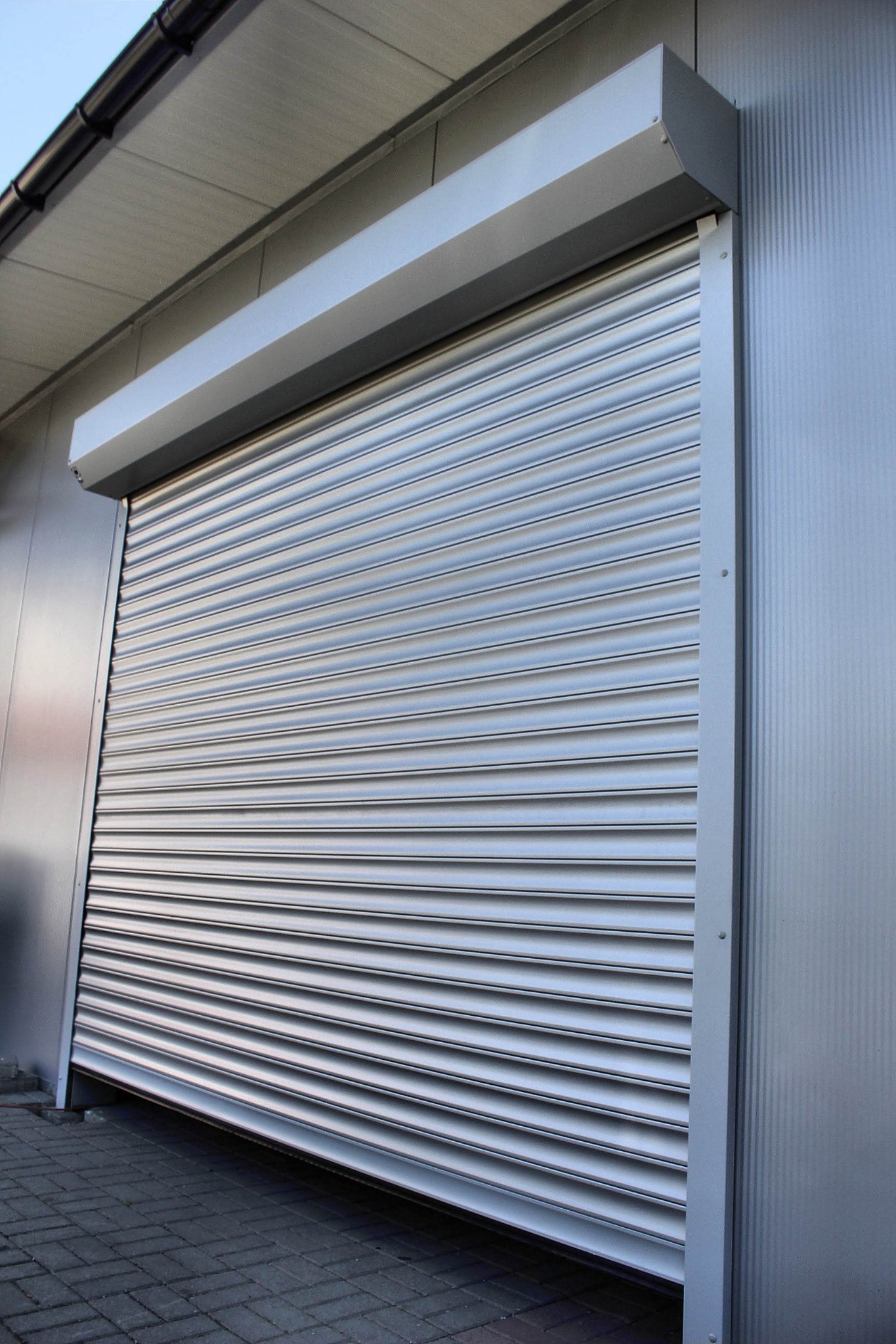 Zamknięta, srebrna brama rolowana garażowa z szarego metalu, widoczna z dołu, na tle szarej ściany budynku i szarej kostki brukowej.
