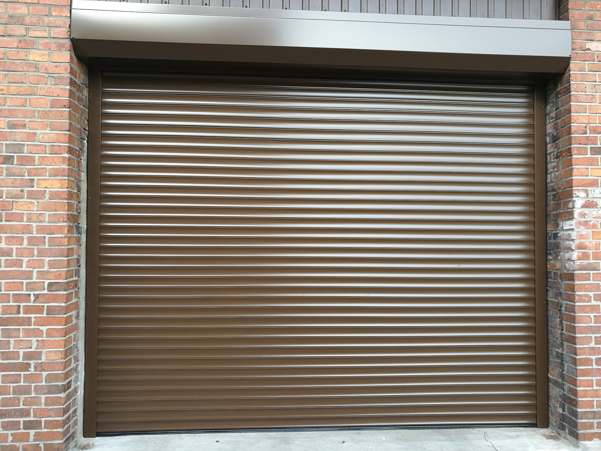Brązowa brama rolowana garażowa zamontowana w murze z cegły, widoczna z bliska, z osłoną nad bramą w tym samym kolorze.
