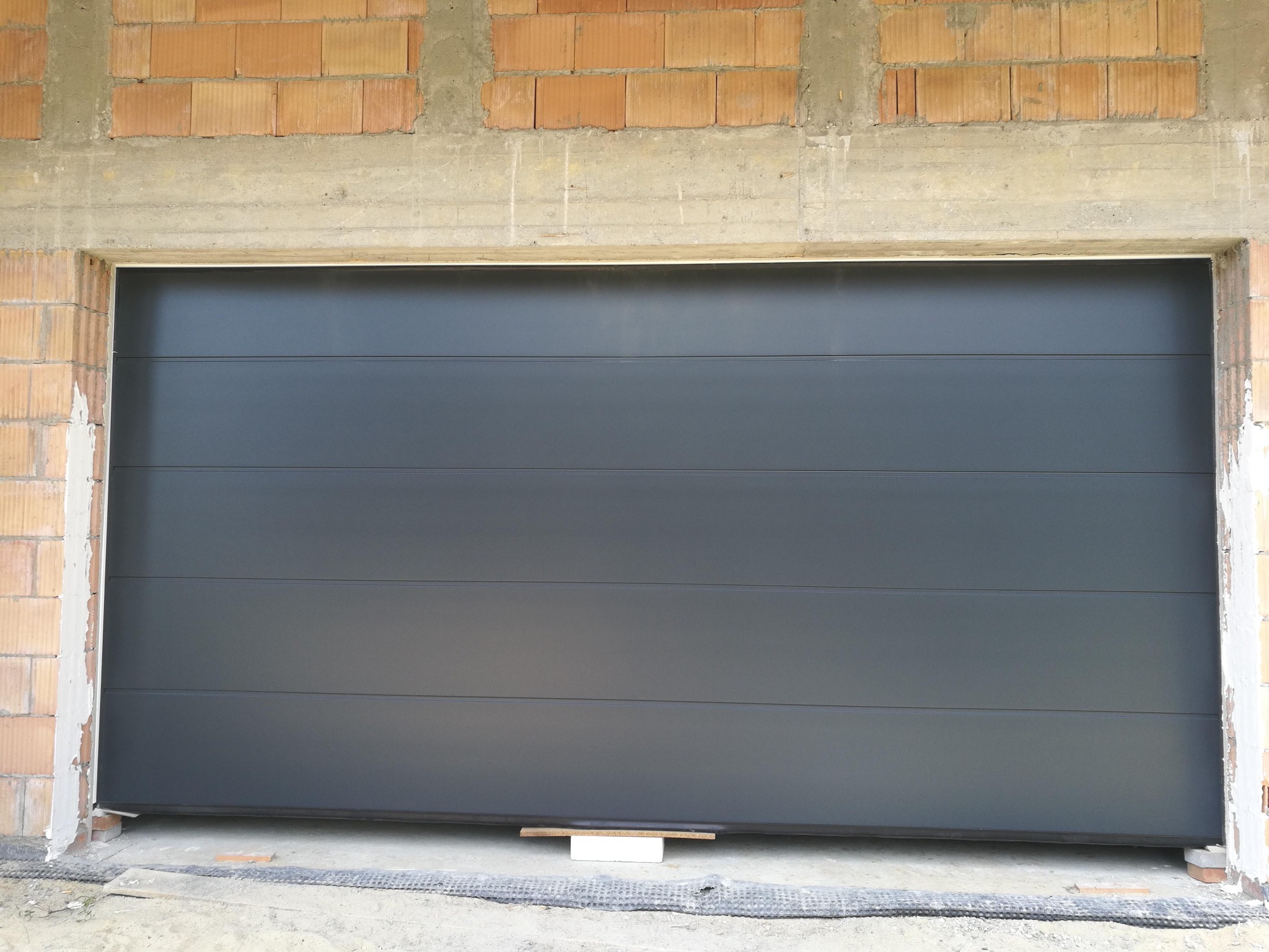 Zamknięta, segmentowa brama w kolorze antracytowym, zamontowana w surowym otworze z cegły i betonu, widoczne detale montażowe.