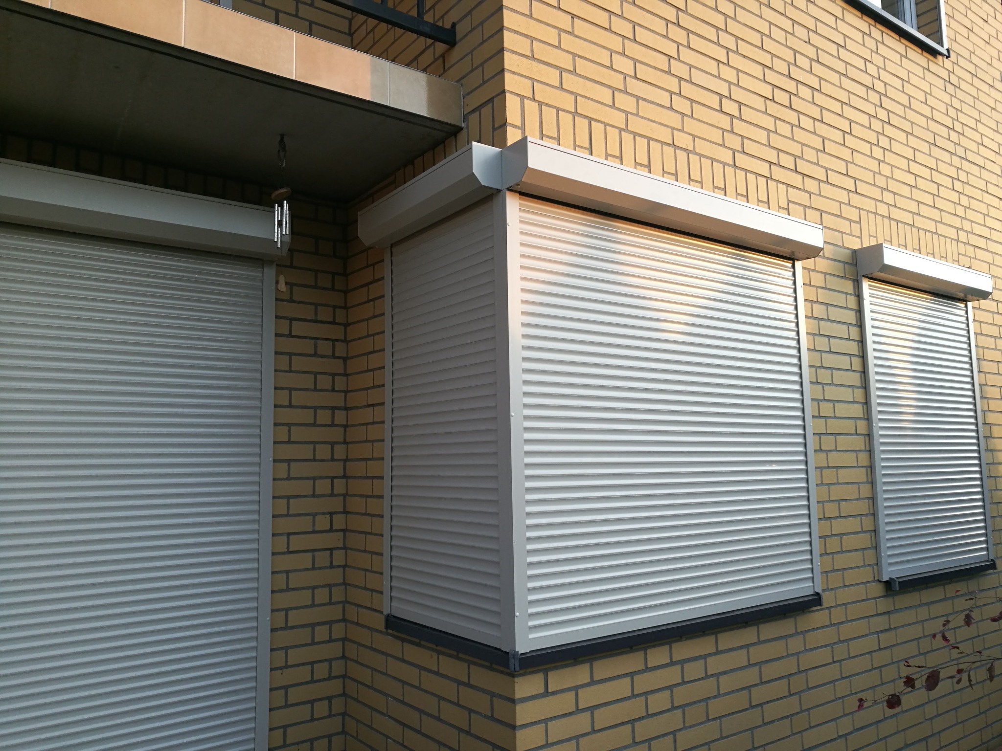 Zewnętrzne, białe rolety aluminiowe na oknach budynku z żółtej cegły, widoczne z boku, z elementami dekoracyjnymi pod dachem.