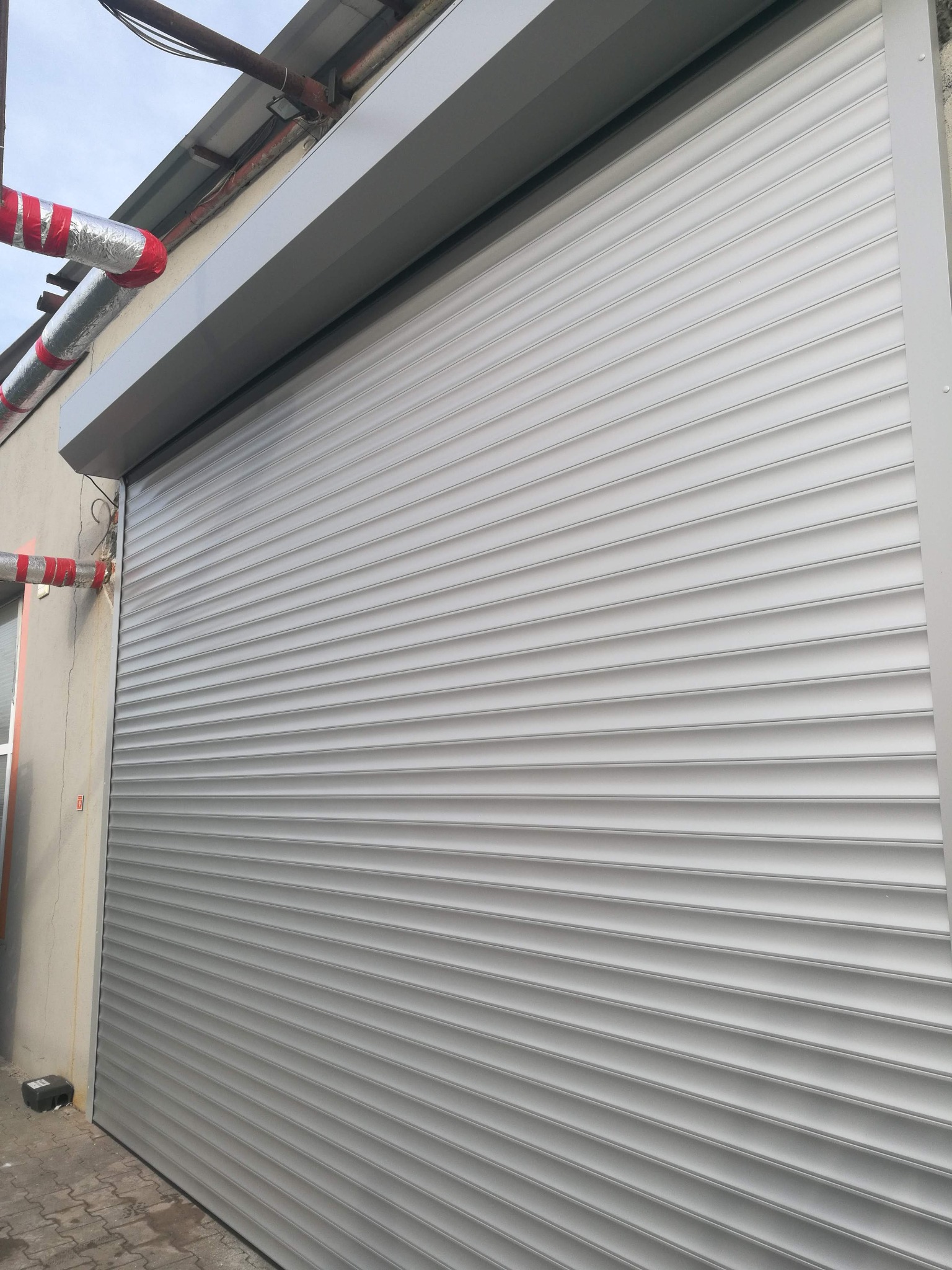 Zamknięta, srebrna brama garażowa z poziomymi panelami, widoczna z boku budynku z rurami grzewczymi owiniętymi taśmą na zewnątrz.